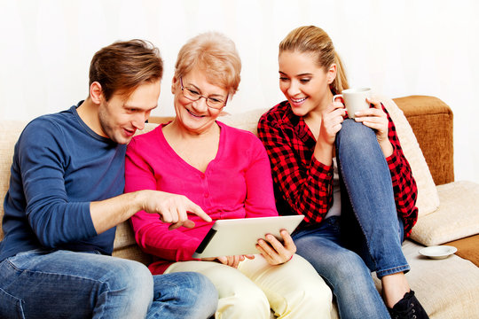 Young couple with old woman sitting on couch and watching something on tablet