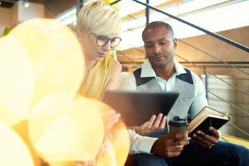 Two creative millenial small business owners working on social media strategy using a digital tablet while sitting in staircase