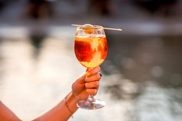 Female hand holding glass with Spritz Aperol alcohol drink decorated with olive on the water chanal...