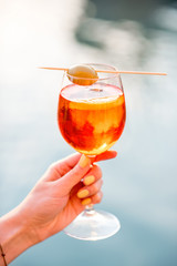 Female hand holding glass with Spritz Aperol alcohol drink decorated with olive on the water chanal...