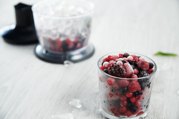 Unfocused blender pot with frozen berries and ice cubes inside behind focused glass with frozen berries Preparation of tasty smoothie step by step process