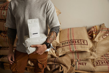 Tattooed barista holds blank package bags with freshly baked coffee beans ready for sale and delivery Big bags on europalet in warehouse behind Professional alternative roasting and brewing
