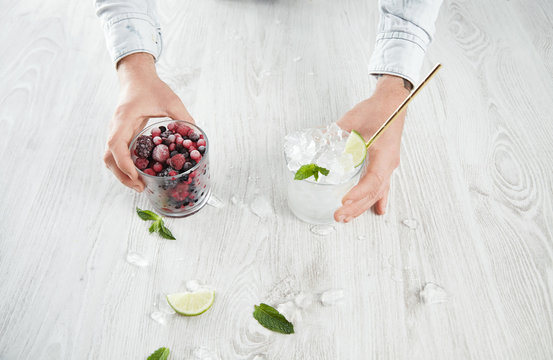 Top view man hands hold glasses with frozen berries and ice cubes with lime, melted crashed ice around, isolated on white wooden table, cocktail set for summer time refreshment