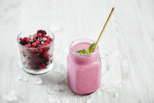 Transparent rustic jar with tasty healthly freshly made pale violet smoothie with drinking straw inside, near unfocused glass with frozen berries Melted ice around isolated on white table