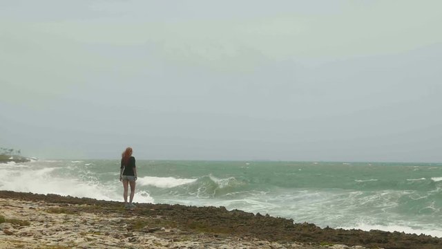 Attractive young woman with long red hair standing on the beach near the storm sea, slow-motion, wide angle