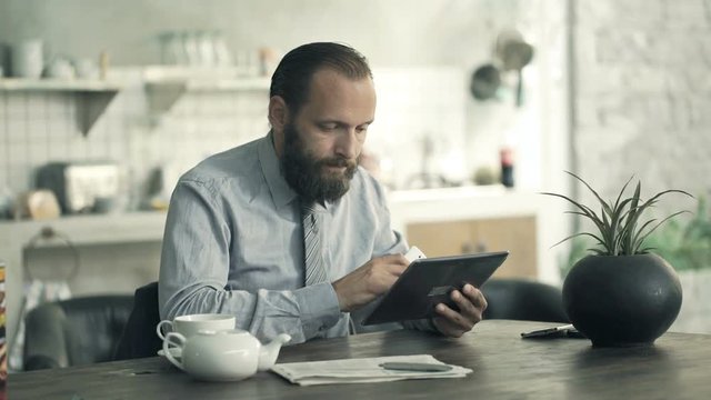 Young businessman comparing data on tablet and smartphone by table in kitchen

