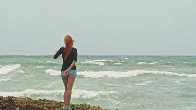 Seductive girl with long red hair standing on the beach near the storm sea, slow-motion