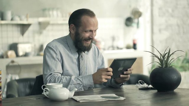 Young, happy businessman playing game on tablet sitting in kitchen 
