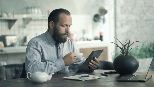 Businessman reading news on tablet and drinking coffee by table in kitchen
