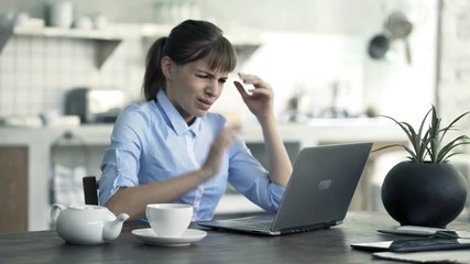 Angry, sad businesswoman working on laptop by table in kitchen at home
