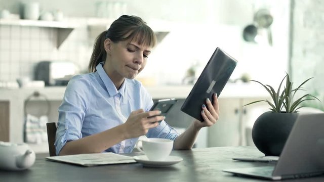 Young businesswoman comparing data on tablet and smartphone by table at home
