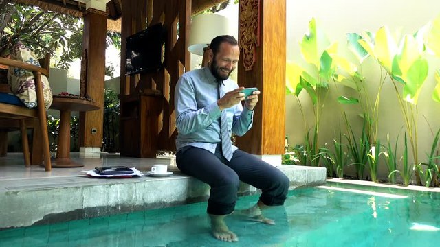 Young businessman playing game sitting on the edge of swimming pool, 4K
