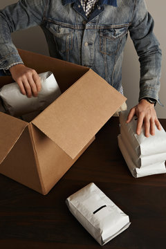 Bearded brutal man in jeans work jacket puts blank sealed hermetic packages inside big carton paper box on wooden table. Special delivery, retail shipping post box top view 