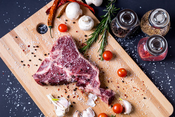 Raw T-bone steak with salt, spices and vegetables on a cutting board, top view.