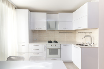 White glossy modern kitchen with stone countertop and built in household appliances in light colors interior