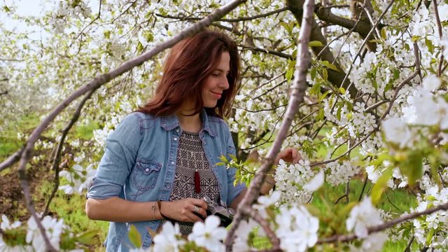 Beautiful young woman smelling and  taking photos with old film camera of blooming tree. Close up. Slow motion