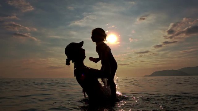 Silhouette of Woman with Toddler in Arms in Sea against Sunset