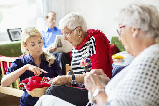 Female caretaker assisting senior women in knitting while man reading book in background at nursing home - Powered by Adobe