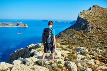 Fototapeta premium male traveler with backpack runs on the cliff against sea and blue sky at early morning. Balos beach on background, Crete, Greece