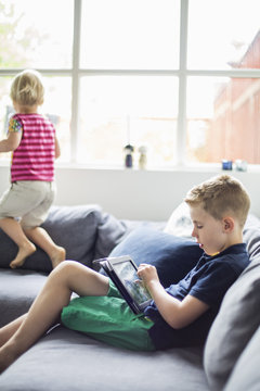 Boy using digital tablet on sofa with sister playing in background