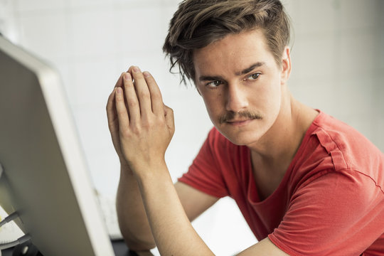 Thoughtful young businessman with hands clasped sitting at computer desk in new office