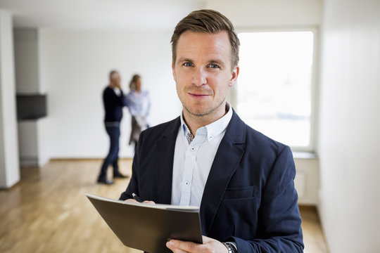 Portrait of confident real estate agent with couple standing in background at home
