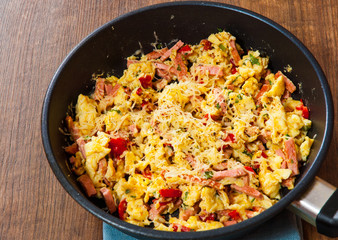 scrambled eggs with ham, vegetables and cheese in a frying pan on wooden table