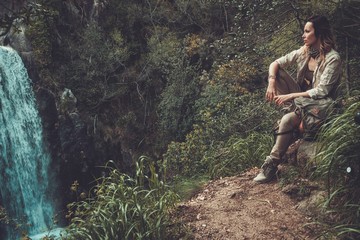 Beautiful woman hiker sitting near waterfall in deep forest.