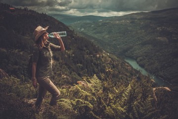 Woman hiker with backpack drinking fresh water from a bottle on a top of mountain.