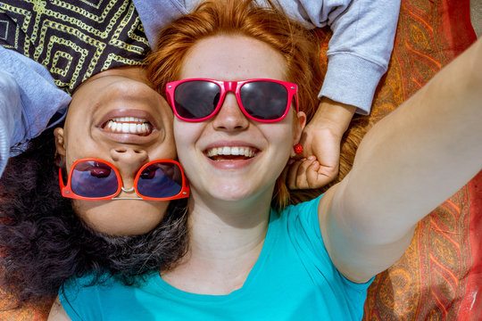 smiling girls multi-cultural best friends lying down take selfie photo wearing eyewear
