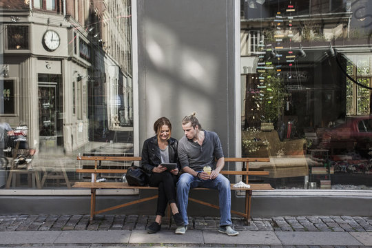 Full length of couple using digital tablet together on sidewalk bench