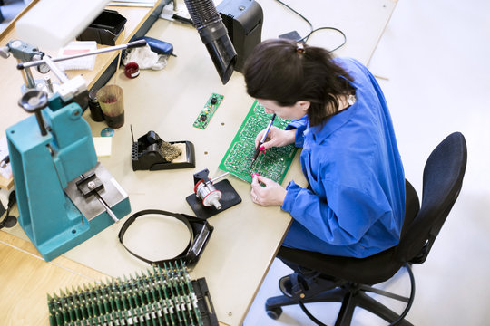 High angle view of female technician soldering circuit board at desk in factory