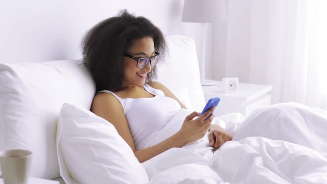 african woman with smartphone in bed at home