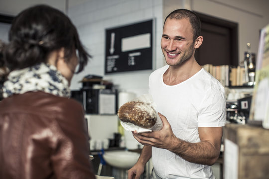 Happy young male owner giving bread loaf to customer in bakery