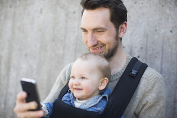 Young man text messaging on mobile phone while carrying baby boy in carrier