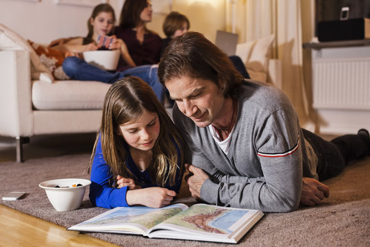 Father and daughter reading map on floor with family sitting on sofa in living room