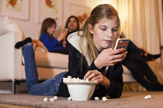Girl using mobile phone while having popcorn on floor with family in background