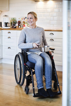 Happy disabled woman in wheelchair looking away while holding saucepan at kitchen