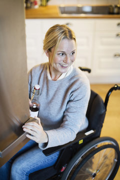 Happy disabled woman with bottle looking away while closing refrigerator's door at kitchen