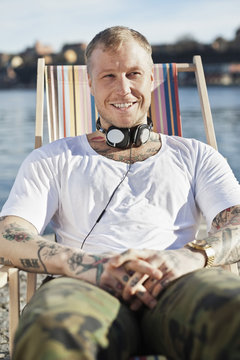 Portrait of a tattooed man with headphones sitting in deck chair at beach