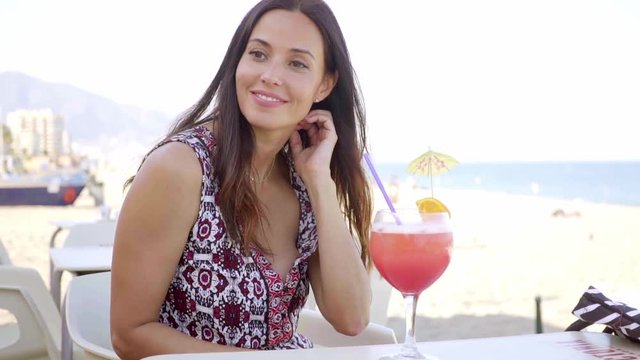 Gorgeous young woman enjoying a tropical cocktail sipping it through a straw as she stands overlooking a beach smiling at the camera