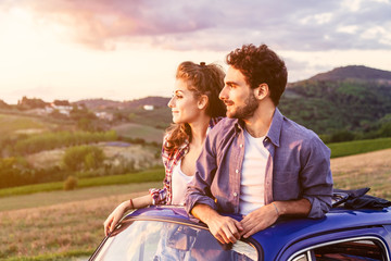 A loving couple, on a summer afternoon, watching sunset from the roof of an old car, around the...