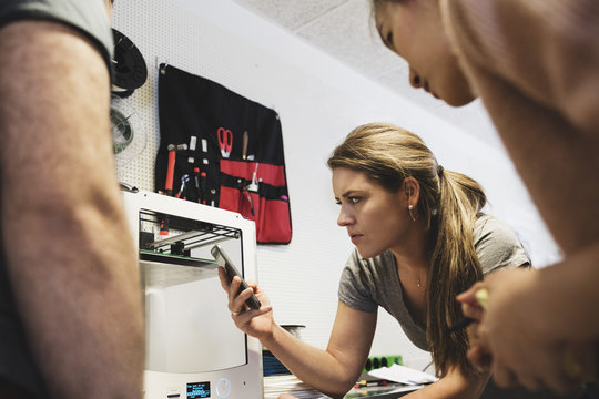 Female engineer holding smart phone and looking at 3D printer with colleagues