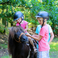 Young children enjoying horse back riding activity. Group of school age kids taking care about their pony and learning how to feed the animal. Summer camp for active holidays.