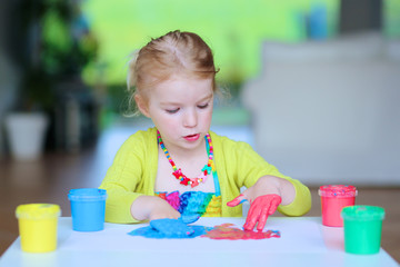 Laughing little child, blonde artistic toddler girl painting and drawing with colorful finger paints indoors at bright room at home or kindergarten.