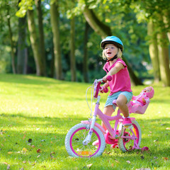 Little child riding her bicycle in the park. Cute preschooler girl learning to cycle with stabilisers wheels. Sportive kid enjoying sunny summer, spring or autumn day outdoors.
