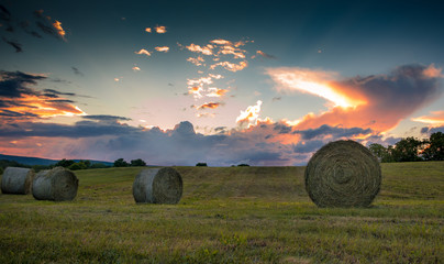 Freshly rolled bales of hay are seen on a rolling hill during a vivid sunset