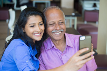 Closeup portrait happy elderly gentleman in pink shirt and lady in blue top taking selfie together, isolated indoors background. Say cheese and smile