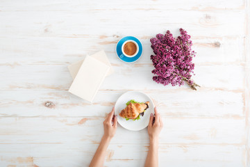 Hands of woman eating croissant and drinking coffee on table