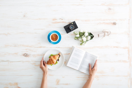 Hands of woman eating croissant with coffee and reading book